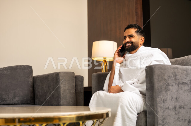 اجراء مكالمة هاتفية عبر الهاتف المحمول, رجل مسلم سعودي خليجي عربي يرتدي ملابس الاحرام يجلس على كنبة في بهو الفندق يمسك بيده الهاتف المحمول, الدليل الالكتروني للحاج و المعتمر, استخدام تطبيقات الجوال للتواصل مع العائلة والأصدقاء