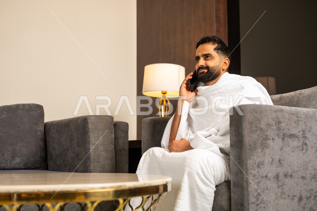 اجراء مكالمة هاتفية عبر الهاتف المحمول, رجل مسلم سعودي خليجي عربي يرتدي ملابس الاحرام يجلس على كنبة في بهو الفندق يمسك بيده الهاتف المحمول, الدليل الالكتروني للحاج و المعتمر, استخدام تطبيقات الجوال للتواصل مع العائلة والأصدقاء