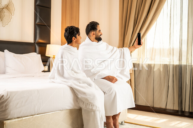 التقاط صور سيلفي باستخدام الهاتف المحمول، أداء مناسك الحج والعمرة، زيارة مكان ومعلم اسلامي ديني مقدس في المملكة العربية السعودية, العبادة والتقرب إلى الله تعالى،  رجل عربي خليجي سعودي يرتدي لباس الاحرام يجلس مع ابنه على سرير الغرفة في الفندق