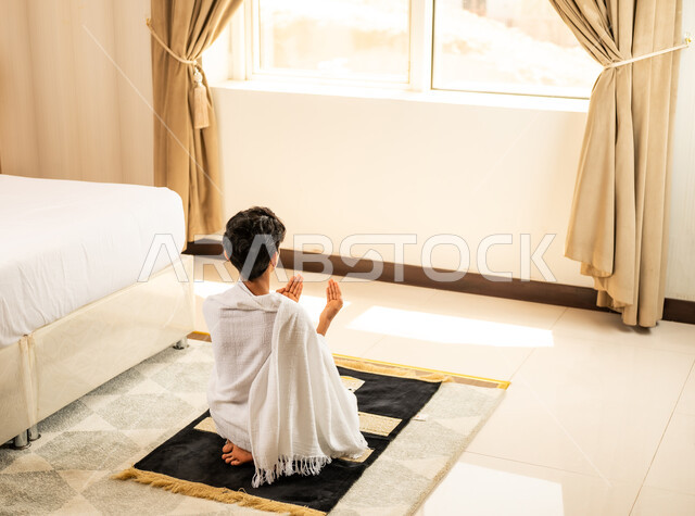 Performing prayers on time, worship and drawing closer to God Almighty, a picture from the back of a Saudi Arabian Gulf boy wearing Ihram clothes sitting in his hotel room with expressions of supplication and prayer, performing the Hajj and Umrah rituals, a sacred Islamic religious place and landmark in the Kingdom of Saudi Arabia