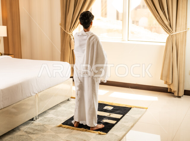 اداء الصلاة على وقتها، العبادة والتقرب إلى الله تعالى، صورة من الخلف لولد عربي خليجي سعودي يرتدي ثياب الاحرام يجلس في غرفته الفندقية بتعابير الدعاء والتضرع, أداء مناسك الحج والعمرة، مكان ومعلم اسلامي ديني مقدس في المملكة العربية السعودية