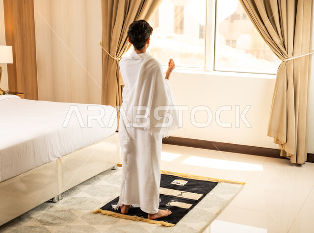اداء الصلاة على وقتها، العبادة والتقرب إلى الله تعالى، صورة من الخلف لولد عربي خليجي سعودي يرتدي ثياب الاحرام يجلس في غرفته الفندقية بتعابير الدعاء والتضرع, أداء مناسك الحج والعمرة، مكان ومعلم اسلامي ديني مقدس في المملكة العربية السعودية
