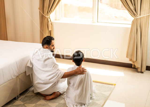 Performing prayers on time, worship and getting closer to God Almighty, a picture from the back of a Saudi Arabian Gulf father wearing Ihram clothes sitting in his hotel room praying and supplicating with his son with expressions of supplication and supplication, performing the Hajj and Umrah rituals, a sacred Islamic religious place and landmark in Saudi Arabia