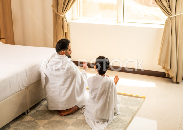 اداء الصلاة على وقتها، العبادة والتقرب إلى الله تعالى، صورة من الخلف لأب عربي خليجي سعودي يرتدي ثياب الاحرام يجلس في غرفته الفندقية يقوم بالدعاء والصلاة برفقة ابنه بتعابير الدعاء والتضرع, أداء مناسك الحج والعمرة، مكان ومعلم اسلامي ديني مقدس في السعودية