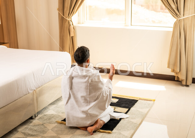 اداء الصلاة على وقتها، العبادة والتقرب إلى الله تعالى، صورة من الخلف لرجل يرتدي ثياب الاحرام يجلس في غرفته الفندقية بتعابير الدعاء والتضرع, أداء مناسك الحج والعمرة، مكان ومعلم اسلامي ديني مقدس في المملكة العربية السعودية