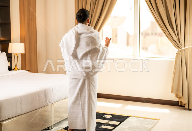 اداء الصلاة على وقتها، العبادة والتقرب إلى الله تعالى، صورة من الخلف لرجل يرتدي ثياب الاحرام يجلس في غرفته الفندقية بتعابير الدعاء والتضرع, أداء مناسك الحج والعمرة، مكان ومعلم اسلامي ديني مقدس في المملكة العربية السعودية