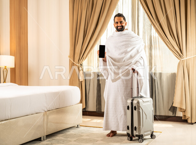 مكان ومعلم اسلامي ديني مقدس في المملكة العربية السعودية, العبادة والتقرب إلى الله تعالى، رجل عربي خليجي سعودي مبتسم يرتدي لباس الاحرام بمسك بيده حقيبة الامتعة يعرض شاشة سوداء فا رغة على الهاتف المحمول, تعابير السرور والسعادة، أداء مناسك الحج والعمرة