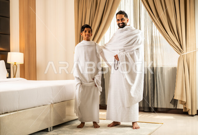 الاستعداد لأداء مناسك الحج والعمرة بأجواء عائلية، العبادة والتقرب إلى الله تعالى، رجل عربي خليجي سعودي يرتدي ثياب الاحرام يقف بجانب ابنه في غرفة الفندق, مكان ومعلم اسلامي ديني مقدس في المملكة العربية السعودية، تعابير الخشوع والتضرع