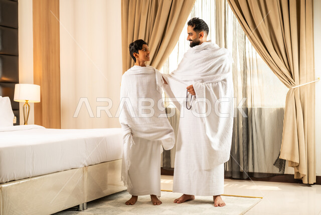 الاستعداد لأداء مناسك الحج والعمرة بأجواء عائلية، العبادة والتقرب إلى الله تعالى، رجل عربي خليجي سعودي يرتدي ثياب الاحرام يقف بجانب ابنه في غرفة الفندق, مكان ومعلم اسلامي ديني مقدس في المملكة العربية السعودية، تعابير الخشوع والتضرع