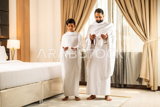 الاستعداد لأداء مناسك الحج والعمرة بأجواء عائلية، العبادة والتقرب إلى الله تعالى، رجل عربي خليجي سعودي يرتدي ثياب الاحرام يقف بجانب ابنه في غرفة الفندق, مكان ومعلم اسلامي ديني مقدس في المملكة العربية السعودية، تعابير الخشوع والتضرع