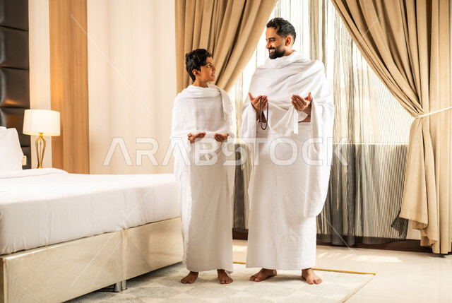الاستعداد لأداء مناسك الحج والعمرة بأجواء عائلية، العبادة والتقرب إلى الله تعالى، رجل عربي خليجي سعودي يرتدي ثياب الاحرام يقف بجانب ابنه في غرفة الفندق, مكان ومعلم اسلامي ديني مقدس في المملكة العربية السعودية، تعابير الخشوع والتضرع