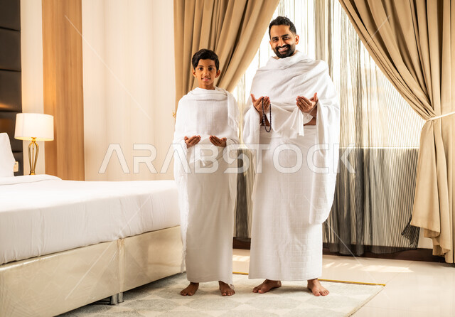 الاستعداد لأداء مناسك الحج والعمرة بأجواء عائلية، العبادة والتقرب إلى الله تعالى، رجل عربي خليجي سعودي يرتدي ثياب الاحرام يقف بجانب ابنه في غرفة الفندق, مكان ومعلم اسلامي ديني مقدس في المملكة العربية السعودية، تعابير الخشوع والتضرع
