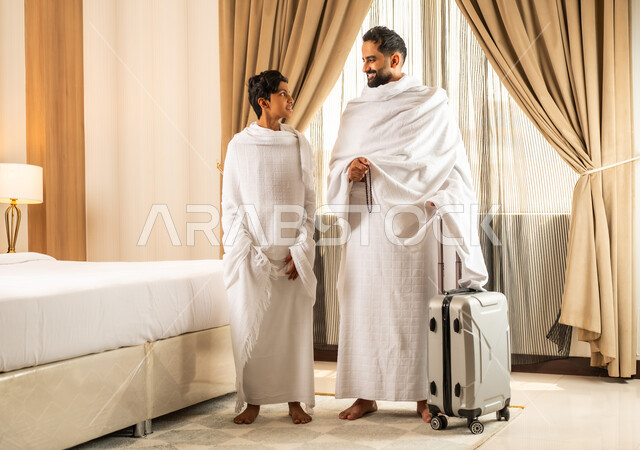 الاستعداد لأداء مناسك الحج والعمرة بأجواء عائلية، العبادة والتقرب إلى الله تعالى، رجل عربي خليجي سعودي يرتدي ثياب الاحرام يقف بجانب ابنه في غرفة الفندق, مكان ومعلم اسلامي ديني مقدس في المملكة العربية السعودية، تعابير الخشوع والتضرع