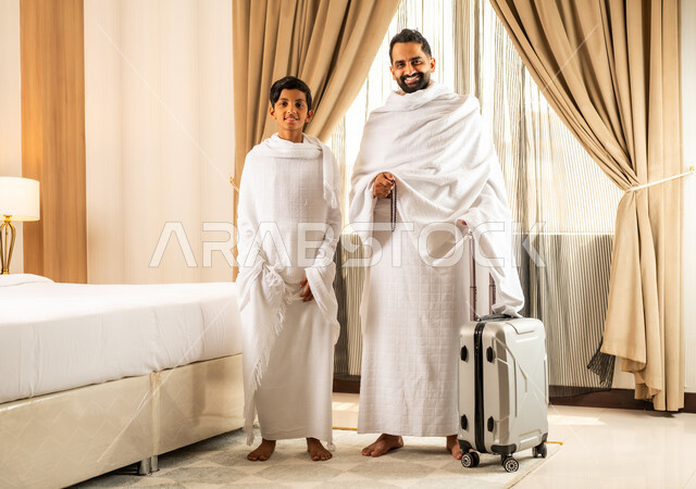 الاستعداد لأداء مناسك الحج والعمرة بأجواء عائلية، العبادة والتقرب إلى الله تعالى، رجل عربي خليجي سعودي يرتدي ثياب الاحرام يقف بجانب ابنه في غرفة الفندق, مكان ومعلم اسلامي ديني مقدس في المملكة العربية السعودية، تعابير الخشوع والتضرع
