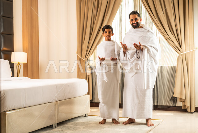 الاستعداد لأداء مناسك الحج والعمرة بأجواء عائلية، العبادة والتقرب إلى الله تعالى، رجل عربي خليجي سعودي يرتدي ثياب الاحرام يقف بجانب ابنه في غرفة الفندق, مكان ومعلم اسلامي ديني مقدس في المملكة العربية السعودية، تعابير الخشوع والتضرع
