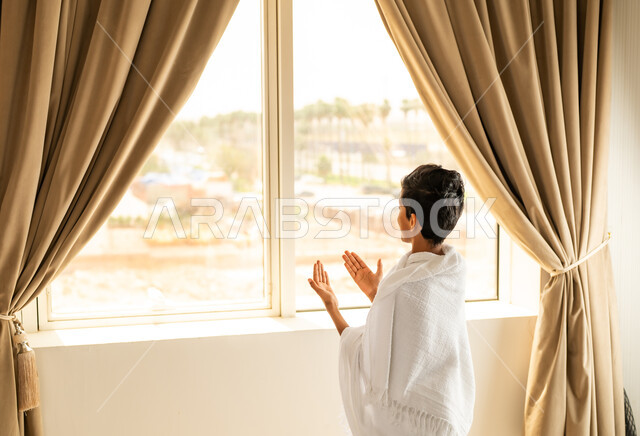 قراءة وتلاوة القرآن الكريم، العبادة والتقرب إلى الله تعالى، الاطلالة على جبال مكة المكرمة، صورة من الخلف لشاب عربي خليجي سعودي يرتدي ثياب الاحرام يمسك بيده المصحف الشريف, أداء مناسك الحج والعمرة، مكان ومعلم اسلامي ديني مقدس في المملكة العربية السعودية