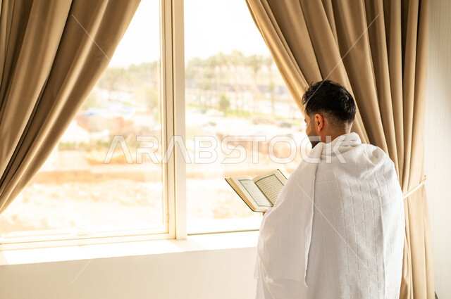 قراءة وتلاوة القرآن الكريم، العبادة والتقرب إلى الله تعالى، الاطلالة على جبال مكة المكرمة، صورة من الخلف لشاب عربي خليجي سعودي يرتدي ثياب الاحرام يمسك بيده المصحف الشريف, أداء مناسك الحج والعمرة، مكان ومعلم اسلامي ديني مقدس في المملكة العربية السعودية