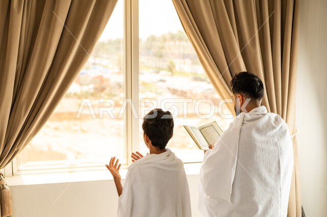 مكان ومعلم اسلامي ديني مقدس في المملكة العربية السعودية, العبادة والتقرب إلى الله تعالى، الاطلالة على جبال مكة المكرمة، صورة من الخلف لرجل عربي خليجي سعودي يرتدي لباس الاحرام يقوم بقراءة القرآن مع ابنه, مقصد المسلمين من أنحاء العالم، أداء مناسك الحج والعمرة