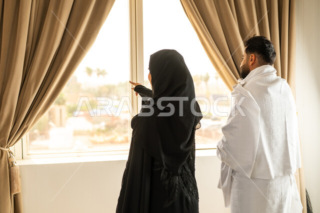 الاطلالة على جبال مكة المكرمة، صورة من الخلف لرجل عربي خليجي سعودي يرتدي لباس الاحرام يقف على واجهه زجاجية مع زوجته, مقصد المسلمين من أنحاء العالم، أداء مناسك الحج والعمرة، مكان ومعلم اسلامي ديني مقدس في المملكة العربية السعودية, العبادة والتقرب إلى الله تعالى