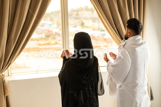 الاطلالة على جبال مكة المكرمة، صورة من الخلف لرجل عربي خليجي سعودي يرتدي لباس الاحرام يقف على واجهه زجاجية مع زوجته, مقصد المسلمين من أنحاء العالم، أداء مناسك الحج والعمرة، مكان ومعلم اسلامي ديني مقدس في المملكة العربية السعودية, العبادة والتقرب إلى الله تعالى