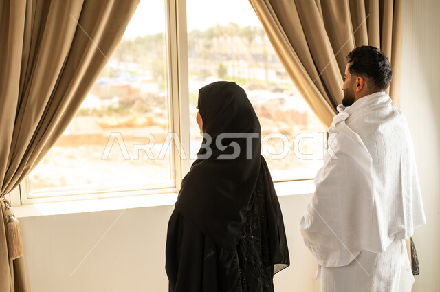 الاطلالة على جبال مكة المكرمة، صورة من الخلف لرجل عربي خليجي سعودي يرتدي لباس الاحرام يقف على واجهه زجاجية مع زوجته, مقصد المسلمين من أنحاء العالم، أداء مناسك الحج والعمرة، مكان ومعلم اسلامي ديني مقدس في المملكة العربية السعودية, العبادة والتقرب إلى الله تعالى