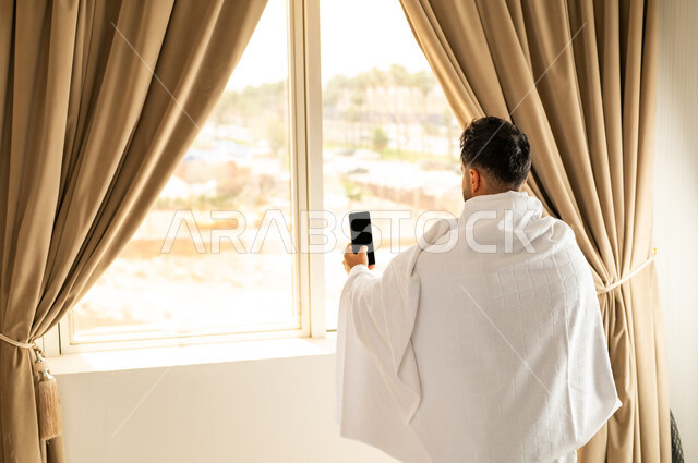 العبادة والتقرب إلى الله تعالى، الاطلالة على ساحة الحرم المكي الشريف، صورة مقربة من الخلف لرجل يرتدي ثياب الاحرام يقف امام واجهه زجاجية ويرفع يديه بتعابير الدعاء والتضرع, أداء مناسك الحج والعمرة، مكان ومعلم اسلامي ديني مقدس في المملكة العربية السعودية