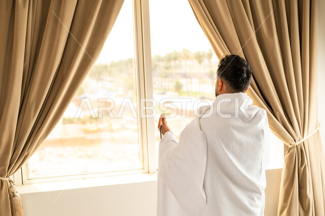 العبادة والتقرب إلى الله تعالى، الاطلالة على ساحة الحرم المكي الشريف، صورة مقربة من الخلف لرجل يرتدي ثياب الاحرام يقف امام واجهه زجاجية ويرفع يديه بتعابير الدعاء والتضرع, أداء مناسك الحج والعمرة، مكان ومعلم اسلامي ديني مقدس في المملكة العربية السعودية