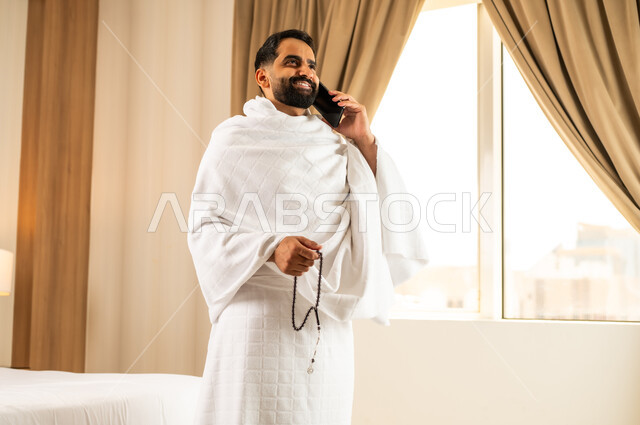 إجراء مكالمه هاتفيه عبر الجوال, رجل مسلم خليجي عربي سعودي يرتدي ملابس الاحرام  يقف في غرفه فندقيه امام الحرم المكي الشريف ، أداء الصلاة ومناسك الحج والعمره في المسجد الحرام في مكة المكرمه، موسم الحج في شهر ذو الحجه