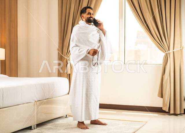 إجراء مكالمه هاتفيه عبر الجوال, رجل مسلم خليجي عربي سعودي يرتدي ملابس الاحرام  يقف في غرفه فندقيه امام الحرم المكي الشريف ، أداء الصلاة ومناسك الحج والعمره في المسجد الحرام في مكة المكرمه، موسم الحج في شهر ذو الحجه