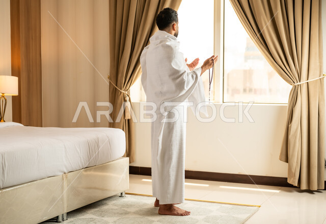 Performing Hajj and Umrah rituals, a sacred Islamic religious place and landmark in the Kingdom of Saudi Arabia, worship and drawing closer to God Almighty, overlooking the courtyard of the Grand Mosque in Mecca, a man wearing Ihram clothes standing in front of a glass facade and raising his hands with expressions of supplication and prayer