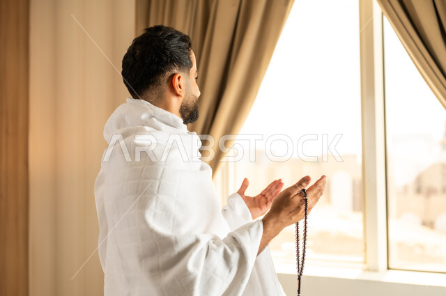 العبادة والتقرب إلى الله تعالى، الاطلالة على ساحة الحرم المكي الشريف، صورة مقربة من الخلف لرجل يرتدي ثياب الاحرام يقف امام واجهه زجاجية ويرفع يديه بتعابير الدعاء والتضرع, أداء مناسك الحج والعمرة، مكان ومعلم اسلامي ديني مقدس في المملكة العربية السعودية