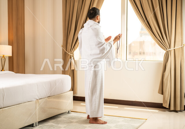أداء مناسك الحج والعمرة، مكان ومعلم اسلامي ديني مقدس في المملكة العربية السعودية، العبادة والتقرب إلى الله تعالى، الاطلالة على ساحة الحرم المكي الشريف، رجل يرتدي ثياب الاحرام يقف امام واجهه زجاجية ويرفع يديه بتعابير الدعاء والتضرع