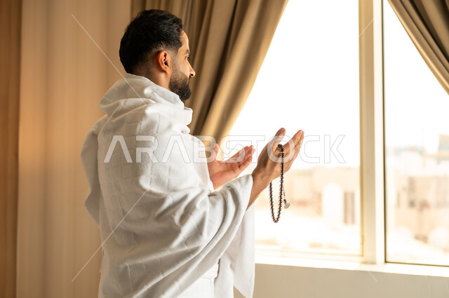 العبادة والتقرب إلى الله تعالى، الاطلالة على ساحة الحرم المكي الشريف، صورة مقربة من الخلف لرجل يرتدي ثياب الاحرام يقف امام واجهه زجاجية ويرفع يديه بتعابير الدعاء والتضرع, أداء مناسك الحج والعمرة، مكان ومعلم اسلامي ديني مقدس في المملكة العربية السعودية