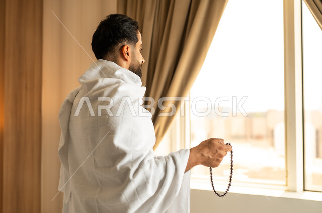Worship and drawing closer to God Almighty, overlooking the courtyard of the Grand Mosque in Mecca, a close-up photo from the back of a man wearing ihram clothes standing in front of a glass facade and raising his hands with expressions of supplication and prayer, performing the Hajj and Umrah rituals, a sacred Islamic religious place and landmark in the Kingdom of Saudi Arabia