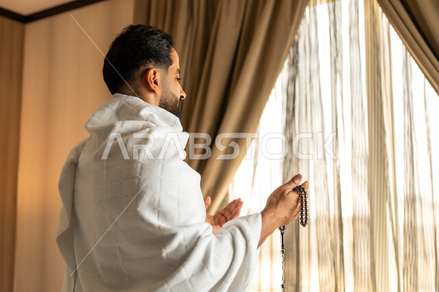 العبادة والتقرب إلى الله تعالى، الاطلالة على ساحة الحرم المكي الشريف، صورة مقربة من الخلف لرجل يرتدي ثياب الاحرام يقف امام واجهه زجاجية ويرفع يديه بتعابير الدعاء والتضرع, أداء مناسك الحج والعمرة، مكان ومعلم اسلامي ديني مقدس في المملكة العربية السعودية