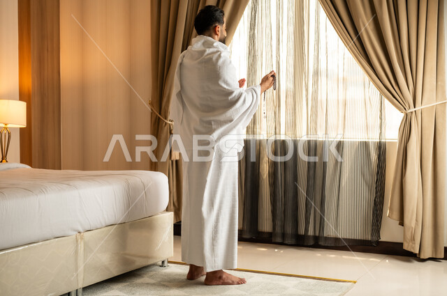 Performing Hajj and Umrah rituals, a sacred Islamic religious place and landmark in the Kingdom of Saudi Arabia, worship and drawing closer to God Almighty, overlooking the courtyard of the Grand Mosque in Mecca, a man wearing Ihram clothes standing in front of a glass facade and raising his hands with expressions of supplication and prayer