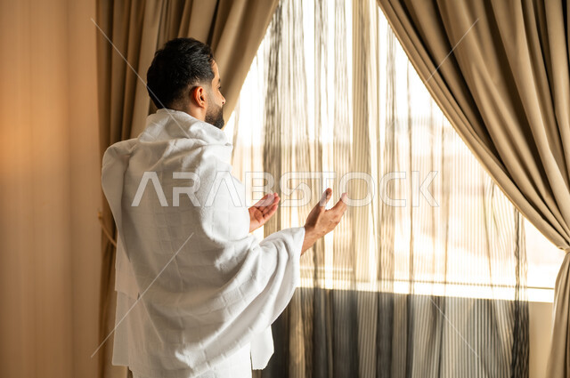 العبادة والتقرب إلى الله تعالى، الاطلالة على ساحة الحرم المكي الشريف، صورة مقربة من الخلف لرجل يرتدي ثياب الاحرام يقف امام واجهه زجاجية ويرفع يديه بتعابير الدعاء والتضرع, أداء مناسك الحج والعمرة، مكان ومعلم اسلامي ديني مقدس في المملكة العربية السعودية