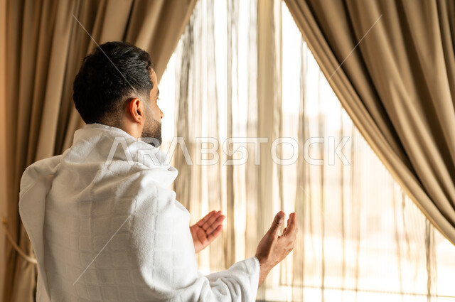 العبادة والتقرب إلى الله تعالى، الاطلالة على ساحة الحرم المكي الشريف، صورة مقربة من الخلف لرجل يرتدي ثياب الاحرام يقف امام واجهه زجاجية ويرفع يديه بتعابير الدعاء والتضرع, أداء مناسك الحج والعمرة، مكان ومعلم اسلامي ديني مقدس في المملكة العربية السعودية