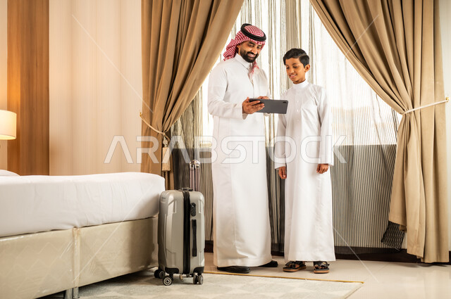 مشاركة الوقت والاهتمامات مع الأبناء، التعلم عن بعد، رجل عربي خليجي سعودي يرتدي الشماغ والثوب التقليدي يدرب ابنه على التعلم من خلال جهاز التابلت المحمول داخل غرفتهما الفندقيه, متابعة الهوايات و النشاطات من خارج المنزل، مكالمات فيديو اونلاين، أجواء عائلية سعيدة