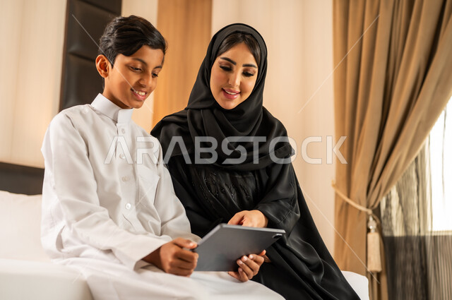 المشاركه بقراءه القصص المفيده مع الأبناء، مفهوم التربيه بالحب والحنان، امرأة عربية خليجية سعودية ترتدي العباءه والحجاب تقرأ الروايات المسليه لابنها في غرفه الفندق عبر جهاز التابلت, استغلال الاوقات في تعليم وتربيه الاولاد، استخدام جهاز تقني حديث