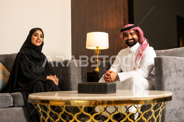 جلسات عائلية سعيدة، زوجان عربيان خليجيان سعوديان يجلسان في غرفة المعيشة يقومان بتبادل الأحاديث والحوارات بإيماءات المودة والتفاهم، الإنصات والاستماع باحترام للحديث بين الزوجين، مفهوم الألفة والمحبة بين الأزواج