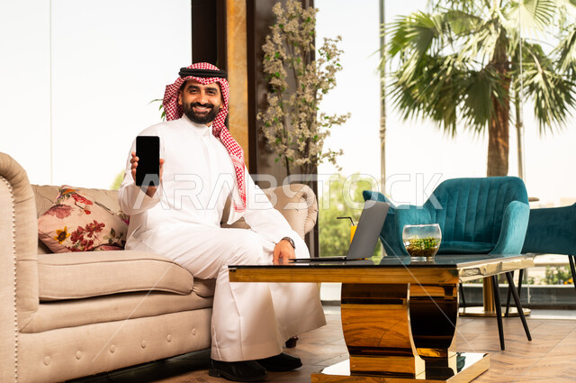عرض شاشة سوداء فارغة عبر الهاتف المحمول، تعابير الاندماج والتركيز أثناء العمل عبر الحاسب المحمول، الجلوس في إحدى الفنادق الراقية، استخدام الأجهزة التكنولوجية الحديثة والمتطورة، رجل عربي خليجي سعودي يرتدي الشماغ والثوب التقليدي يجلس في لوبي فندق يستخدم اللابتوب