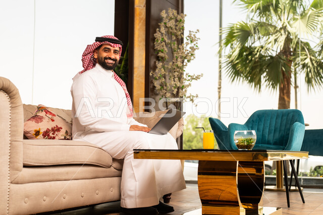 تعابير الاندماج والتركيز أثناء العمل عبر الحاسب المحمول، الجلوس في إحدى الفنادق الراقية، استخدام الأجهزة التكنولوجية الحديثة والمتطورة، رجل عربي خليجي سعودي يرتدي الشماغ والثوب التقليدي يجلس في لوبي فندق يستخدم اللابتوب
