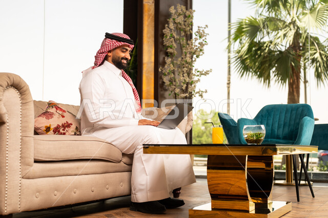 تعابير الاندماج والتركيز أثناء العمل عبر الحاسب المحمول، الجلوس في إحدى الفنادق الراقية، استخدام الأجهزة التكنولوجية الحديثة والمتطورة، رجل عربي خليجي سعودي يرتدي الشماغ والثوب التقليدي يجلس في لوبي فندق يستخدم اللابتوب