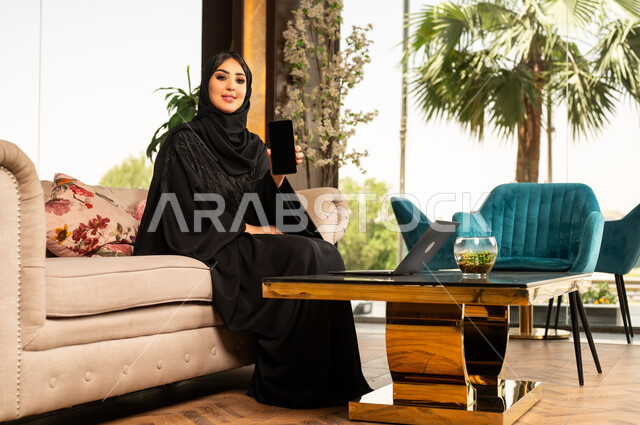 عرض شاشة سوداء فارغة على الهاتف المحمول، استخدام الأجهزة التكنولوجية الحديثة والمتطورة، سيدة أعمال عربية خليجية سعودية مبتسمة ترتدي الحجاب والعباءة تجلس في لوبي فندق فاخر تقوم بالعمل على الحاسوب المحمول، تعابير وايماءات الاندماج والتركيز لإنجاز المهام عن بعد