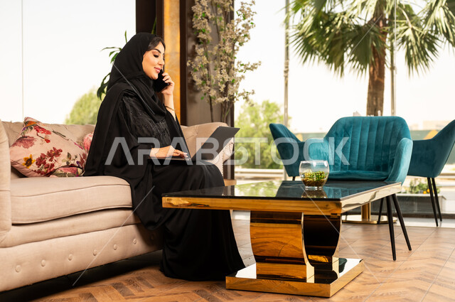 اجراء مكالمة هاتفية عبر الهاتف المحمول، استخدام الأجهزة التكنولوجية الحديثة والمتطورة، سيدة أعمال عربية خليجية سعودية مبتسمة ترتدي الحجاب والعباءة تجلس في لوبي فندق فاخر تقوم بالعمل على الحاسوب المحمول، تعابير وايماءات الاندماج والتركيز لإنجاز المهام عن بعد