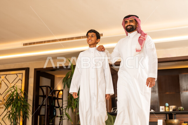 The concept of tourism during summer vacation, spending a pleasant summer trip, preparing to travel on a tourist tour, a Saudi Arabian Gulf father wearing the traditional thobe and shemagh walking around the hotel lobby with his son, gestures of happiness, pleasure and joy
