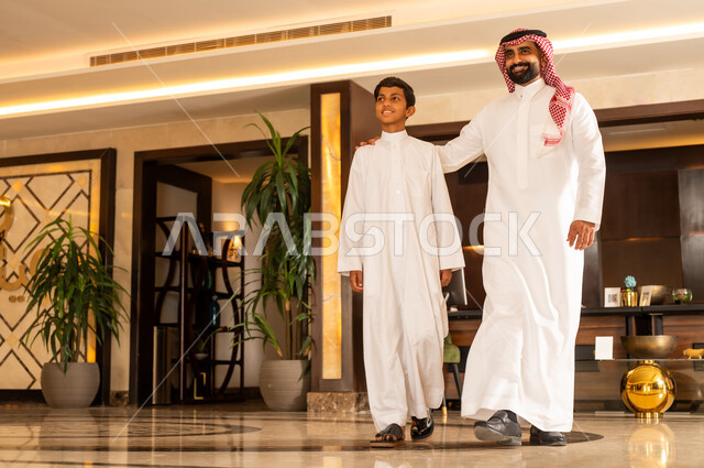 The concept of tourism during summer vacation, spending a pleasant summer trip, preparing to travel on a tourist tour, a Saudi Arabian Gulf father wearing the traditional thobe and shemagh walking around the hotel lobby with his son, gestures of happiness, pleasure and joy