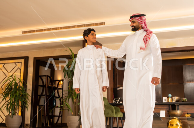 مفهوم السياحة في إجازة الصيف، قضاء رحلة صيفية ممتعة، الاستعداد للسفر في جولة سياحية، أب عربي خليجي سعودي يرتدي الثوب التقليدي والشماغ يتجول في بهو الفندق برفقة ابنه، إيماءات السعادة والسرور والسعادة
