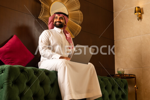 مفهوم اداره الاعمال عن بعد, استخدام التقنيات الحديثه في التواصل مع الاهل والاصدقاء وانجاز الاعمال,  الجلوس في إحدى الفنادق الراقية، رجل عربي خليجي سعودي يرتدي الشماغ والثوب التقليدي يجلس في لوبي الفندق ويعمل على جهاز الحاسوب المحمول